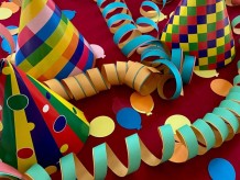 Bunte Luftschlangen, Konfetti in Form von bunten Ballons und bunte Partyhütchen auf einer dunkelroten Fläche.